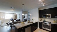This single-floor space showcases a thoughtfully designed open-concept layout that integrates the kitchen, dining, and living areas into one cohesive environment. The kitchen serves as the focal point, featuring dark espresso cabinetry paired with granite countertops that offer ample prep and storage space. A central island with an undermount double sink provides additional workspace and acts as a natural divider between the kitchen and the adjoining living area. Pendant lighting hangs above the island, adding warmth and visual interest, while a track lighting system overhead ensures the space is well-lit throughout. Stainless steel appliances, including a gas range and microwave, complete the modern kitchen aesthetic. Light hardwood-style flooring runs throughout, creating a warm and cohesive base that complements the darker cabinet tones. The open living area beyond the kitchen is bright and airy, with natural light filtering in from windows along the far wall. A glimpse into an adjacent bedroom reveals a mirrored feature that amplifies light and adds depth. The overall ambiance is contemporary and functional, balancing clean lines with comfortable, livable design.