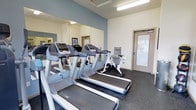 This fitness center is a dedicated, single-floor space designed to support an active lifestyle. The room is outfitted with multiple treadmills and a stationary spin bike, all arranged to maximize usability and flow. Large wall-mounted mirrors line one side of the room, creating a sense of expanded space while allowing users to monitor their form during workouts. The flooring consists of durable, dark rubber gym matting, a practical and resilient surface well-suited for exercise equipment. A built-in water fountain is conveniently positioned along the wall, providing easy hydration access mid-workout. Lighting is provided by overhead fluorescent fixtures, ensuring the space is well-lit throughout. The walls feature a clean, two-tone design with a muted blue accent along the upper portion and neutral white below, contributing to a calm and focused atmosphere. A glazed entry door allows natural light to filter into the space, adding a bright and welcoming quality. The overall design is functional and straightforward, prioritizing equipment accessibility and an open layout that accommodates multiple users simultaneously.