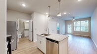 This 1-bedroom, 1-bathroom unit offers a thoughtfully designed layout that maximizes its footprint across a single floor. The open-concept kitchen and living area serves as the heart of the space, anchored by a central island with a granite countertop and an integrated stainless steel sink, illuminated by stylish pendant lights overhead. The kitchen is fully equipped with stainless steel appliances, including a refrigerator, electric range, and over-the-range microwave, complemented by crisp white cabinetry and a tile backsplash. Light hardwood-style flooring flows throughout the main living areas, creating a bright and cohesive aesthetic. The living room offers generous natural light through large windows and a glass door, enhanced by a ceiling fan overhead. A set of double closet doors provides convenient built-in storage. The bedroom features plush carpeting, a ceiling fan, and two windows that bring in natural light. A dedicated closet offers additional storage. The bathroom is clean and functional, complete with a tiled shower-tub combination, white cabinetry, granite countertops, and hardwood-style flooring. Neutral wall tones throughout lend the space a modern, move-in-ready feel.