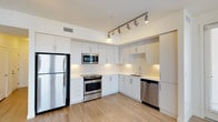 This single-floor residence showcases a contemporary design with clean lines and a bright, open aesthetic throughout. The kitchen is a standout feature, outfitted with a full suite of stainless steel appliances, including a refrigerator, gas range, built-in microwave, and dishwasher. Crisp white cabinetry extends to the ceiling, offering generous storage, while light-toned countertops and a white tile backsplash contribute to the space's airy, modern feel. A track lighting system mounted overhead provides adjustable, focused illumination across the workspace. Light-toned hardwood-style flooring runs throughout, adding warmth and continuity to the interior. A ceiling-mounted ventilation duct ensures efficient airflow. The unit also features a private balcony framed by textured stucco walls, offering expansive views of a resort-style courtyard below. The outdoor area is highlighted by a large, freeform swimming pool, lounge seating, and shade structures, all set against a backdrop of multi-story residential buildings. This outdoor vantage point enhances the living experience, blending indoor comfort with a vibrant community atmosphere.