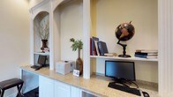 This single-floor space presents a thoughtfully designed workspace that combines functionality with classic architectural detailing. The standout feature of the area is its custom built-in workstation, which showcases a generous granite countertop providing ample surface area for computer equipment and office essentials. White cabinetry beneath the countertop offers convenient storage while maintaining a clean, polished aesthetic.

Elegant arched alcoves framed with white molding are integrated into the design, serving as both decorative and functional display niches. These architectural details lend the space a refined, traditional character. The warm, neutral wall tones complement the crisp white millwork throughout.

The workspace is equipped with multiple computer monitors and a printer, making it well-suited for productivity. Decorative accents, including a classic world globe on a turned pedestal, potted greenery, and curated books, add warmth and personality to the environment. A sculptural branch arrangement in a vase contributes an organic touch to the overall composition.

The overall ambiance is organized and inviting, striking a balance between a professional work environment and a comfortable, homey aesthetic. The careful integration of architectural details and functional furnishings makes this a versatile and well-considered space.