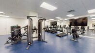 Located at 901 N Market Street in Wilmington, Delaware, this fitness center is a comprehensively outfitted exercise facility designed to support a wide range of workout routines. The space is organized into distinct functional zones across a single floor, creating a logical and efficient layout for users.

The cardio area features a row of True-brand treadmills and elliptical machines, each equipped with individual display screens, arranged along a wall of large mirrors that visually expand the space. Bright, recessed panel lighting set within a drop ceiling provides consistent, even illumination throughout. The distinctive blue speckled rubber flooring runs throughout the entire facility, offering both durability and a clean, professional aesthetic.

A dedicated strength and cable machine section houses a multi-function cable station and additional resistance equipment, positioned to allow ample space for safe use. A separate open area accommodates functional training and free weight exercises, with a well-organized dumbbell rack, medicine balls, and a wall-mounted rig for suspension training. Rowing machines and an air bike are also available in this zone.

The facility features white walls, large mirrored panels that enhance the sense of openness, and glass partition walls in certain areas. The overall ambiance is clean, modern, and well-maintained, reflecting a professional fitness environment.