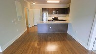 This single-floor space is designed with a clean, modern aesthetic that emphasizes openness and functionality. The heart of the home is the open-concept kitchen, which features rich, dark espresso cabinetry that extends to the upper walls, providing generous storage. A large kitchen island anchors the space, topped with sleek granite countertops that offer ample workspace and a built-in sink. Stainless steel appliances — including a refrigerator, electric range, and over-the-range microwave — lend a polished, contemporary feel. A mosaic tile backsplash adds a subtle decorative accent along the cooking area. Warm hardwood flooring flows seamlessly throughout the open living area, reflecting natural and overhead light to create an inviting ambiance. Crown molding details along the ceiling add an elegant finishing touch. A hallway visible from the main living area leads to additional rooms, hinting at a thoughtful, well-organized floor plan. The neutral wall tones complement the darker fixtures and flooring, resulting in a balanced, welcoming interior. Overall, the space blends practical design with contemporary style, making it both visually appealing and highly livable.