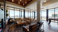 Located near I-25 in Denver, Colorado, this single-floor amenity space offers an impressive blend of contemporary design and elevated urban living. The space is defined by expansive floor-to-ceiling windows that wrap around the main lounge area, flooding the interior with natural light and providing sweeping panoramic views of the Denver skyline and surrounding landscape.

The main lounge features soaring ceilings with a striking wood-paneled section outfitted with pendant lighting, adding warmth and texture to the open layout. A decorative mosaic column serves as an architectural focal point, complementing the rich hardwood flooring found throughout. Multiple seating arrangements—including sectional sofas, tufted leather benches, and classic leather armchairs—create distinct yet connected gathering zones. A fireplace with a sculptural wood accent wall anchors one end of the lounge, adding a dramatic design element alongside a wall-mounted display screen.

The kitchen area is outfitted with stainless steel appliances, granite countertops, and a stunning iridescent mosaic tile backsplash framed by warm wood cabinetry. A separate game room provides a dedicated entertainment space with a poker table, track lighting, and views to the exterior. Together, these spaces create a sophisticated, versatile environment designed for both social gatherings and relaxed everyday use.