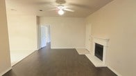 This single-floor home offers a well-designed layout centered around a generous living area that serves as the heart of the space. The living room is highlighted by a classic white-mantled fireplace, which serves as a focal point and adds a timeless, welcoming character to the room. Dark hardwood flooring runs throughout, providing a rich contrast against the neutral, light-toned walls and creating a warm, grounded ambiance. A ceiling fan with an integrated light fixture is centrally mounted, ensuring both comfort and adequate illumination across the open space. The room benefits from a clean, open feel, with doorways leading to additional areas of the home, suggesting a practical and connected floor plan. Transition areas between rooms feature lighter flooring, subtly delineating different functional zones within the space. The overall aesthetic is understated and neutral, offering a versatile canvas well-suited for a variety of interior styles. The single-floor configuration ensures ease of movement throughout, making this a highly accessible and functional living environment.