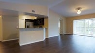 This single-floor space showcases a thoughtfully designed open-concept layout that connects the kitchen and living areas in a seamless, functional flow. The kitchen is a standout feature, equipped with stainless steel appliances — including a refrigerator and built-in microwave — complemented by rich dark cabinetry and a granite-topped breakfast bar that provides both a casual dining option and a natural visual divider between the kitchen and the main living space. The warm beige walls provide a neutral backdrop that enhances the richness of the dark hardwood flooring, which runs consistently throughout the open areas, lending the space a cohesive and polished aesthetic. Vaulted or elevated ceilings add a sense of openness and volume, while strategically placed HVAC vents indicate year-round climate control. Natural light filters in through windows visible in the background, brightening the living area and creating an inviting atmosphere. A hallway visible beyond the main living space suggests additional rooms, adding to the functional depth of the layout. Overall, this space balances modern finishes with a warm, livable ambiance.