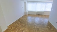 This single-floor space presents a clean, minimalist interior highlighted by classic parquet hardwood flooring laid in a geometric basket-weave pattern, adding warmth and visual texture throughout. The open layout provides a generous sense of space, with crisp white walls that enhance brightness and create a neutral canvas adaptable to a variety of uses. A large window fitted with horizontal blinds allows ample natural light to fill the room, contributing to an airy, welcoming atmosphere. A baseboard heating unit sits beneath the window, offering a practical heating solution integrated seamlessly into the design. Subtle architectural detailing, including a small recessed ledge along one wall, adds modest dimension to the otherwise straightforward layout. The absence of interior partitions in the main area creates an open, flexible environment. The overall ambiance is understated and functional, with the parquet flooring serving as the primary design feature, lending a timeless, classic character to the space. The combination of natural light, hardwood flooring, and white walls gives the interior a bright, neutral, and well-maintained feel.