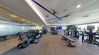 This modern fitness center offers a spacious, single-floor layout thoughtfully designed to accommodate a comprehensive workout experience. The space features a distinctive tray ceiling with bold navy blue accent lines and integrated recessed lighting, contributing to a polished, contemporary ambiance. Rubber flooring spans the entire area, providing both durability and safety for a variety of exercises.

The cardio section is a standout feature, lined with Matrix treadmills and elliptical trainers positioned along a wall of floor-to-ceiling windows. These large windows flood the space with natural light and offer expansive outdoor views of the surrounding greenery and skyline, creating an energizing atmosphere during workouts.

The strength training area is equally well-appointed, housing a multi-station cable machine, a rack of dumbbells ranging across multiple weights, adjustable benches, and a variety of resistance machines. A large flat-screen television is mounted on a mirrored wall, and full-length mirrors throughout the space enhance both functionality and the sense of openness.

A striking dark accent wall featuring a cosmic-themed mural adds a creative visual element, while glass partitions and ceiling fans contribute to both the aesthetic and ventilation of the space. The overall design balances athletic functionality with refined interior detailing.