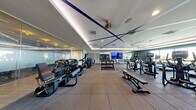 This modern fitness center offers a spacious, single-floor layout thoughtfully designed to accommodate a comprehensive workout experience. The space features a distinctive tray ceiling with bold navy blue accent lines and integrated recessed lighting, contributing to a polished, contemporary ambiance. Rubber flooring spans the entire area, providing both durability and safety for a variety of exercises.

The cardio section is a standout feature, lined with Matrix treadmills and elliptical trainers positioned along a wall of floor-to-ceiling windows. These large windows flood the space with natural light and offer expansive outdoor views of the surrounding greenery and skyline, creating an energizing atmosphere during workouts.

The strength training area is equally well-appointed, housing a multi-station cable machine, a rack of dumbbells ranging across multiple weights, adjustable benches, and a variety of resistance machines. A large flat-screen television is mounted on a mirrored wall, and full-length mirrors throughout the space enhance both functionality and the sense of openness.

A striking dark accent wall featuring a cosmic-themed mural adds a creative visual element, while glass partitions and ceiling fans contribute to both the aesthetic and ventilation of the space. The overall design balances athletic functionality with refined interior detailing.