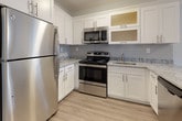 Located at 107 Wartburg Ave in Copiague, NY, this single-floor unit offers a thoughtfully designed open-concept layout that seamlessly connects the living area and kitchen. The kitchen is a standout feature, showcasing white shaker-style cabinetry with frosted glass upper panels, granite-look countertops, a classic subway tile backsplash, and a full suite of stainless steel appliances including a refrigerator, range, and over-the-range microwave. A peninsula with matching countertop provides additional prep space and defines the kitchen from the expansive living area. Light wood-tone luxury vinyl plank flooring runs throughout, lending warmth and cohesion to the entire space. Multiple windows invite natural light into both the living area and the bedrooms, offering views of the surrounding neighborhood. The bedrooms are finished in soft, neutral blue-gray tones and feature sliding closet doors with built-in rod storage. Baseboard heating is present throughout. A dedicated laundry closet houses a stacked washer and dryer unit for added convenience. Clean white trim, neutral walls, and contemporary finishes throughout give the space a fresh, move-in-ready character.