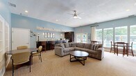 Located at 9440 Pleasant Point Way in Ashland, Virginia, this single-floor community space offers a well-appointed layout comprising an open living room and a full kitchen. The living room is a welcoming gathering area highlighted by a striking floor-to-ceiling stacked stone fireplace with a mounted flat-screen television, serving as the room's central focal point. Soft blue walls, neutral carpeting, and a ceiling fan with integrated lighting contribute to a relaxed, comfortable atmosphere. Ample seating includes a large sectional sofa and additional dining-height tables and chairs, making the space suitable for both casual lounging and social gatherings. Large windows and glass patio doors flood the room with natural light and offer views of the outdoor pool area, enhancing the open and airy feel. French doors connect to a secondary seating nook, adding flexibility to the layout. The kitchen features rich, dark cabinetry paired with granite countertops and a peninsula with bar seating. Tile flooring, recessed lighting, and pendant fixtures complete the functional yet stylish space. Together, the living room and kitchen create a cohesive, community-centered environment ideal for residents to gather, socialize, and relax.