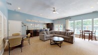 Located at 9440 Pleasant Point Way in Ashland, Virginia, this single-floor community space offers a well-appointed layout comprising an open living room and a full kitchen. The living room is a welcoming gathering area highlighted by a striking floor-to-ceiling stacked stone fireplace with a mounted flat-screen television, serving as the room's central focal point. Soft blue walls, neutral carpeting, and a ceiling fan with integrated lighting contribute to a relaxed, comfortable atmosphere. Ample seating includes a large sectional sofa and additional dining-height tables and chairs, making the space suitable for both casual lounging and social gatherings. Large windows and glass patio doors flood the room with natural light and offer views of the outdoor pool area, enhancing the open and airy feel. French doors connect to a secondary seating nook, adding flexibility to the layout. The kitchen features rich, dark cabinetry paired with granite countertops and a peninsula with bar seating. Tile flooring, recessed lighting, and pendant fixtures complete the functional yet stylish space. Together, the living room and kitchen create a cohesive, community-centered environment ideal for residents to gather, socialize, and relax.