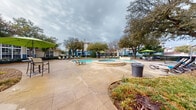 This outdoor amenity area serves as a central gathering space for the residential community, combining recreational and relaxation features in a cohesive, well-maintained environment. The centerpiece is a generously sized swimming pool with clear blue water, complemented by an adjacent raised spa finished in classic stone coping. The expansive concrete sundeck surrounds both the pool and spa, providing ample space for lounge chairs and small patio tables shaded by vibrant green umbrellas — ideal for outdoor leisure.

Mature trees are thoughtfully positioned throughout the grounds, offering natural shade and a sense of established landscaping. The pool area is enclosed by a black iron fence, providing both security and a defined boundary for the space.

Adjacent to the pool deck is a covered outdoor lounge area framed by decorative arched columns in a neutral stucco finish with blue accent bases. This covered patio is furnished with cushioned seating and a coffee table, creating a comfortable shaded retreat. Large windows and French doors connect this porch to the interior clubhouse building, which features a traditional architectural style with a pitched roof and white-trimmed façade. Together, these elements create a resort-style outdoor environment designed for community gathering and relaxation.