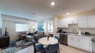 Located at 1012 Shiloh Way in Morrisville, NC, this single-floor suite offers a thoughtfully designed layout that balances comfort and functionality. The open-concept living and dining area serves as the heart of the space, featuring light wood-style flooring, recessed lighting, and large windows fitted with horizontal blinds that allow for ample natural light. The living area is furnished with a plush sofa, accent chairs, and a mint-green ottoman, complemented by vibrant teal abstract artwork that sets a cohesive color palette throughout.

The full kitchen is equipped with stainless steel appliances — including a range, microwave, refrigerator, and dishwasher — alongside white cabinetry, granite countertops, and a double sink, making it well-suited for extended stays or everyday cooking.

Two bedrooms each feature a king-sized bed, dedicated storage cabinetry, nightstands, and matching teal wall art. One bedroom includes an en-suite bathroom with a glass-enclosed shower, while the other connects to a full bathroom with a shower-tub combination. A stacked washer and dryer unit is conveniently integrated into the bathroom area, adding a practical amenity for longer-term occupants. The overall aesthetic is clean, modern, and inviting throughout.
