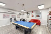Located at 65 Wynford Heights Cres in Toronto, this dedicated recreational room is a thoughtfully designed amenity space built for leisure and socializing. The room is anchored by a full-size pool table with a vivid blue felt surface, positioned at the center and illuminated by a stylish trio of pendant lights suspended from chains above — a standout design feature that sets the tone for the space.

The flooring throughout is light-toned laminate wood, lending a clean and modern aesthetic that complements the bright white walls. A wall-mounted split air conditioning unit ensures year-round comfort. Recessed skylight-style flush ceiling panels provide consistent, even lighting across the room.

The lounge area features a bold red leather sofa paired with framed mirrors and curated wall art, creating a vibrant and inviting atmosphere. A glass-top dining table with black chairs offers a secondary gathering spot, suitable for board games or casual conversation — a chess set is notably present. A wall-mounted rack neatly stores pool cues, while a small television on a stand adds to the entertainment options.

Decorative framed artwork, including London-themed photography prints and abstract pieces, adds personality to the space. The room is accessible via a dedicated door with glazed panels, and the overall layout is open and functional, making it an ideal communal amenity for residents.