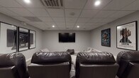 Located at 459 23rd St in Oakland, CA, this single-floor screening room is designed to deliver a true cinema experience. The space is finished with neutral gray walls and a drop ceiling fitted with recessed lighting and a centrally mounted HVAC cassette unit, creating a controlled, comfortable atmosphere ideal for extended viewing sessions.

At the heart of the room is a wall-mounted flat-screen television serving as the primary display. Seating is thoughtfully layered to accommodate various preferences: a row of dark leather home theater recliners with built-in cup holders offers a classic cinema feel, while an assortment of large, plush bean bag chairs provides a more relaxed, casual option. Subtle raised platform details along the walls add architectural dimension to the room.

The décor embraces a cinematic theme, with framed movie posters — including Black Panther, Trainspotting, and Moneyball — displayed prominently on the walls, alongside a striking triptych of large-format photography prints depicting a vintage camera. A dedicated in-ceiling speaker system supports quality audio throughout the space.

Practical features include carpeted tile flooring for sound absorption, a secured exit door with emergency signage, and a wall-mounted access control panel. The overall ambiance is sleek, moody, and purpose-built for film enthusiasts.
