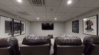 Located at 459 23rd St in Oakland, CA, this single-floor screening room is designed to deliver a true cinema experience. The space is finished with neutral gray walls and a drop ceiling fitted with recessed lighting and a centrally mounted HVAC cassette unit, creating a controlled, comfortable atmosphere ideal for extended viewing sessions.

At the heart of the room is a wall-mounted flat-screen television serving as the primary display. Seating is thoughtfully layered to accommodate various preferences: a row of dark leather home theater recliners with built-in cup holders offers a classic cinema feel, while an assortment of large, plush bean bag chairs provides a more relaxed, casual option. Subtle raised platform details along the walls add architectural dimension to the room.

The décor embraces a cinematic theme, with framed movie posters — including Black Panther, Trainspotting, and Moneyball — displayed prominently on the walls, alongside a striking triptych of large-format photography prints depicting a vintage camera. A dedicated in-ceiling speaker system supports quality audio throughout the space.

Practical features include carpeted tile flooring for sound absorption, a secured exit door with emergency signage, and a wall-mounted access control panel. The overall ambiance is sleek, moody, and purpose-built for film enthusiasts.