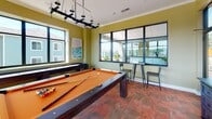 Located at 139 Alexander Bank Dr. in Mooresville, NC, this single-floor game room is designed for social entertainment and leisure. The space centers around a full-size pool table with an orange felt surface, accented by a striking industrial-style pendant light fixture overhead. Warm olive-green walls and richly textured terracotta-toned flooring give the room a vibrant, welcoming character, while crown molding adds a polished architectural detail.

Large black-framed windows line the walls, flooding the room with natural light and offering open views of the surrounding community and lush greenery. A shuffleboard table is positioned along one wall beneath the windows, complementing the billiards setup. A wall-mounted flat-screen TV and a dedicated pool cue rack provide both entertainment and practicality. A dark wood credenza offers additional storage, while industrial-style bar-height tables and chairs create casual seating areas throughout.

Original artwork, including a coastal landscape painting, adds a decorative touch that enhances the room's relaxed ambiance. Access to an adjacent outdoor area is visible through a glass-paneled door, further extending the social atmosphere of the space. Overall, this game room strikes a balance between energetic recreation and comfortable, well-appointed design.
