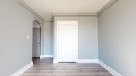 This single-floor unit offers a clean, modern aesthetic defined by neutral gray walls, crisp white trim, and light hardwood-style flooring that flows consistently throughout the space. The entry area features a solid front door, a utility panel, and access to an interior room, establishing a practical and organized layout from the moment you step inside.

The main living areas are bright and open, benefiting from multiple double-hung windows that invite natural light and offer views of the surrounding neighborhood. The rooms are well-proportioned, providing a comfortable sense of space without feeling overwhelming. Each room is finished with wall-mounted heating units, ensuring year-round comfort.

The kitchenette is efficiently designed, featuring dark wood cabinetry, upper storage, a stainless steel sink, and a compact electric range with an oven — ideal for everyday meal preparation. Natural light streams into this area, enhancing its functional appeal.

The bathroom is finished with classic white subway tile surrounding a full soaking tub, complemented by a small frosted window for natural light and ventilation. The clean, timeless design gives the bathroom a fresh and polished look.

Overall, the unit presents a cohesive, move-in-ready aesthetic with consistent flooring, a neutral color palette, and well-maintained finishes throughout — making it a practical and comfortable living space.