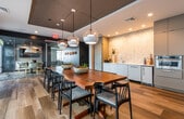 This thoughtfully designed single-floor communal space blends contemporary aesthetics with functional amenities, creating an inviting environment for gathering and relaxation. The layout flows between two distinct zones: a dining and kitchen area, and a lounge and media area.

The dining area centers around a substantial solid-wood communal table paired with mid-century modern chairs, all set atop warm hardwood flooring. Geometric pendant lights hang overhead, while a dramatic two-tone ceiling—featuring a textured dark inset panel—adds architectural depth. Floor-to-ceiling windows flood the space with natural light and provide direct outdoor access.

Adjacent to the dining area, the kitchenette is equipped with sleek light-blue cabinetry, white countertops, a stainless steel sink, a built-in oven, and a refrigerator. A striking hexagonal marble mosaic tile backsplash serves as the kitchen's focal point, extending to the ceiling.

The lounge area transitions to plush carpeted flooring and features generous curved sofas, swivel chairs, and low stone coffee tables arranged around a wall-mounted television. A dramatic highlight of this space is the panoramic floor-to-ceiling windows showcasing breathtaking beachfront and ocean views, making it a standout destination for leisure and entertainment.