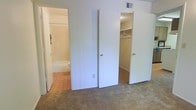 This single-floor residential space offers a practical and functional layout designed for comfortable everyday living. The bedroom area is finished with wall-to-wall carpeting that adds warmth and softness underfoot, creating a cozy atmosphere. Two doorways open from the bedroom — one leading to a spacious walk-in closet outfitted with a hanging rod and ample storage space, ideal for organizing wardrobes and personal belongings. The second doorway provides direct access to a full bathroom, which features a combined bathtub and shower unit along with a toilet, all finished in neutral, light tones that contribute to a clean and bright feel. The bathroom is laid with hard-surface flooring, contrasting the carpeted bedroom for practical functionality. White walls and white-painted door frames throughout maintain a simple, neutral aesthetic that allows for easy personalization. A ceiling vent is visible, indicating a central HVAC system for climate control. The overall design is straightforward and efficient, making this space well-suited for those seeking a no-fuss, move-in-ready residential environment with convenient storage and private bathroom access directly from the sleeping area.
