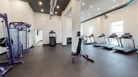 Located at 1728 Port in Rockingham, VA, this single-floor fitness center is thoughtfully designed to accommodate a full range of workout needs. The main exercise area features soaring ceilings with exposed industrial-style ductwork, recessed lighting, and durable rubber flooring, creating an energetic yet functional atmosphere. Large windows along one wall flood the cardio zone with natural light and offer open outdoor views, providing an inviting backdrop for treadmills, ellipticals, stationary bikes, and rowing machines arranged in a well-organized layout. The strength training section is equally well-appointed, housing a full rack of dumbbells, cable machines, a power rack, and adjustable benches — all in a coordinated purple-and-chrome aesthetic that gives the space a modern, professional feel. Full-length mirrors line portions of the walls, enhancing the sense of space and supporting proper form during workouts. A separate studio room, finished with wood-look luxury vinyl plank flooring, a drop ceiling with skylights, and crisp white walls, provides a versatile space ideal for cycling, functional training, or group classes. Medicine balls, a spin bike, and floor mat storage are among the equipment found here. The facility is clean, bright, and well-maintained throughout.