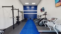 Located at 5736 Remington Cir in Fort Worth, TX, this dedicated fitness facility is designed to support a comprehensive workout experience within a single, open-plan space. The room features soaring ceilings that lend an airy, open feel, complemented by recessed flush lighting panels that ensure bright, even illumination throughout. A bold navy blue accent wall with crisp white horizontal stripes serves as the visual centerpiece, giving the space an energetic and motivating atmosphere. A vibrant blue turf runner stretches the length of the room, dividing the floor and providing a functional lane for agility drills and sled pushes. The flooring consists of durable dark rubber matting, ideal for high-impact training. On the strength side, a full power rack is equipped with TRX suspension trainers, gymnastic rings, a barbell with weight plates, and a flat bench, supported by additional free weights and a sled. The cardio section is equally well-appointed, housing a StairMaster, elliptical, treadmills, an air bike, and a ski erg-style machine. A wall-mounted flat-screen TV is positioned for easy viewing during workouts. Large French doors at one end of the room introduce natural light and provide a welcoming entry point, rounding out this thoughtfully appointed training environment.