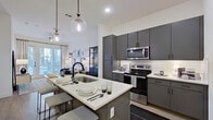 This contemporary 1-bedroom, 1-bathroom residence offers a well-designed layout that maximizes functionality and style across a single floor. The open-concept kitchen is a standout feature, showcasing slate-grey cabinetry extending to the ceiling, a white subway tile backsplash, stainless steel appliances including a refrigerator, electric range, and built-in microwave, and a central island with a white quartz countertop that seats multiple guests beneath elegant globe pendant lights. The kitchen flows seamlessly into the living room, which is bathed in natural light from large floor-to-ceiling windows and a glass-paneled door opening to a balcony. A ceiling fan adds comfort, and light wood-style flooring runs throughout the common areas, enhancing the bright, airy atmosphere. The bedroom is a serene retreat with its own access to natural light through expansive windows, a ceiling fan, and ample space for a full furniture arrangement. The bathroom features a clean, all-white design with a grey floating vanity, a walk-in shower enclosed by black-framed glass doors, and marble-style tile walls and flooring. A dedicated closet completes the thoughtfully organized layout of this modern residence.