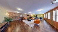 Located at 1632 Aliceanna St in Baltimore's vibrant Fells Point neighborhood, this single-floor communal lounge offers a generously proportioned, open-plan gathering space with a well-curated industrial-modern aesthetic. Rich hardwood flooring runs throughout, anchoring the room's warm yet contemporary character.

Two defining architectural features set the tone: a full exposed brick wall lined with multiple windows that flood the space with natural light, and a striking reclaimed wood accent wall adorned with a decorative geometric panel and bar-height seating — ideal for casual conversation or socializing. The walls are painted in a bold chartreuse-green, adding a lively pop of color that complements the natural textures throughout.

The lounge is thoughtfully divided into distinct activity zones. A central seating area features a grouping of vibrant orange swivel chairs and a leather ottoman atop a striped area rug, centered around a wall-mounted flat-screen TV. Adjacent to this is a dedicated game area complete with a full-size pool table, pendant lighting overhead, and a mounted TV, creating an engaging entertainment hub. A separate lounge nook with navy swivel chairs and a round accent table provides a more relaxed retreat. Recessed and flush-mount ceiling lighting ensures the entire space is well-illuminated throughout the day and evening.