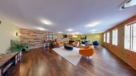 Located at 1632 Aliceanna St in Baltimore's vibrant Fells Point neighborhood, this single-floor communal lounge offers a generously proportioned, open-plan gathering space with a well-curated industrial-modern aesthetic. Rich hardwood flooring runs throughout, anchoring the room's warm yet contemporary character.

Two defining architectural features set the tone: a full exposed brick wall lined with multiple windows that flood the space with natural light, and a striking reclaimed wood accent wall adorned with a decorative geometric panel and bar-height seating — ideal for casual conversation or socializing. The walls are painted in a bold chartreuse-green, adding a lively pop of color that complements the natural textures throughout.

The lounge is thoughtfully divided into distinct activity zones. A central seating area features a grouping of vibrant orange swivel chairs and a leather ottoman atop a striped area rug, centered around a wall-mounted flat-screen TV. Adjacent to this is a dedicated game area complete with a full-size pool table, pendant lighting overhead, and a mounted TV, creating an engaging entertainment hub. A separate lounge nook with navy swivel chairs and a round accent table provides a more relaxed retreat. Recessed and flush-mount ceiling lighting ensures the entire space is well-illuminated throughout the day and evening.
