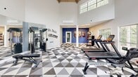 This fitness center in Birmingham, Alabama, is housed in a striking single-room facility that makes a strong architectural impression. The space is defined by its dramatically tall, octagonal-style ceiling with exposed wood detailing at the apex, flanked by a continuous band of clerestory windows that flood the interior with natural light. Large windows and glass double-entry doors along the perimeter further enhance the bright, open atmosphere and provide views of the surrounding greenery.

The floor is covered in a bold geometric carpet pattern of white, gray, and taupe triangles, adding a dynamic visual energy to the space. A structural column near the center anchors the layout while maintaining an open, unobstructed feel throughout.

Equipment is thoughtfully arranged to maximize usability. Cardio machines, including treadmills and a stationary cycle, are positioned along one side, accompanied by a wall-mounted flat-screen television. A dedicated strength-training area features a multi-station cable machine, a weight bench, a dumbbell rack, and a medicine ball and stability ball station. A large wall mirror expands the sense of space and supports exercise form. An accent wall in deep blue, adorned with motivational artwork and a bold "No Pain No Gain" wall decal, adds personality and energy to the overall ambiance. A wall-mounted split HVAC unit ensures year-round comfort.