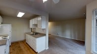 This single-floor space offers an open, functional layout that connects the kitchen and living areas in a straightforward, practical design. The kitchen features white cabinetry — both upper and lower — providing ample storage, complemented by a countertop with an integrated sink and faucet. A skylight positioned above the kitchen draws in natural light, brightening the work area. The hardwood flooring in the kitchen transitions smoothly into carpet in the adjoining living area, creating a clear visual distinction between the two spaces. The living area is generously sized, with neutral-toned walls and carpeting that contribute to a clean, understated aesthetic. A ceiling fan is centrally mounted, offering both air circulation and subtle overhead lighting. A wall-mounted vent ensures climate control throughout the space. The overall design is minimal and practical, making it highly adaptable for various furnishing arrangements. Neutral tones throughout the space create a cohesive, move-in ready environment suited to a range of personal styles.