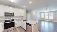 This thoughtfully designed 1-bedroom, 1-bathroom unit offers a functional and modern living experience across a single floor. The open-concept kitchen and living area serves as the heart of the space, featuring crisp white shaker cabinetry, granite countertops, a subway tile backsplash, and a full suite of stainless steel appliances including a refrigerator, range, microwave, and dishwasher. A peninsula island with a built-in sink adds additional prep space and defines the transition into the living area. The living room is bright and airy, benefiting from a large triple-pane window that draws in natural light and a ceiling fan overhead, all set atop rich, dark hardwood-style flooring. The bedroom is a comfortable retreat finished with plush carpeting and a window with outdoor views, while two doors lead conveniently to both the bathroom and a walk-in closet. The bathroom is well-equipped with white cabinetry, granite countertops, a single-basin vanity, and an adjoining walk-in closet with built-in shelving. Neutral warm-gray tones are carried throughout the unit, lending a clean, cohesive, and move-in-ready aesthetic.