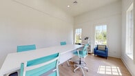 Located at 650 Dekalb St in Auburn, Alabama, this single-room workspace offers a thoughtfully designed environment that maximizes both function and comfort. The main room serves as a versatile living and working area, anchored by a large white conference-style table surrounded by modern teal ergonomic chairs, making it well-suited for meetings or collaborative tasks. Light hardwood flooring runs throughout, complementing the crisp white walls and contributing to the space's clean, professional aesthetic. Notably tall ceilings amplify the sense of openness, preventing the space from feeling confined despite its compact footprint. Multiple large, grid-paned windows line the walls, flooding the interior with abundant natural light and offering pleasant views of the surrounding neighborhood's greenery and streetscape. A seating nook along one wall features two navy blue armchairs flanking a white lattice-front cabinet, providing a comfortable area for informal conversation or a brief respite. A set of elegant glass-panel French doors with transom windows connects the room to the broader building, adding a touch of architectural refinement. Recessed lighting provides supplemental illumination, ensuring the space is well-lit at all times of day. The overall ambiance is bright, airy, and inviting.