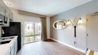 Located at 11260 Point East Dr in Rancho Cordova, California, this single-floor unit offers a thoughtfully arranged layout comprising one bedroom, two bathrooms, a kitchen, and a living room. The living area features light wood-look flooring, textured accent walls, and a large sliding glass door dressed with neutral drape curtains that invite natural light and offer views of surrounding palm trees and the neighborhood beyond. A decorative oval mirror and wall-mounted lighting add character to the space. The kitchen is efficiently designed with gray shaker-style cabinetry extending to the ceiling for ample storage, white countertops, a smooth cooktop, stainless steel appliances including a microwave and refrigerator, and a single-basin sink. An arched passageway provides an architectural detail that connects spaces with a classic touch. The spacious primary bedroom is finished with patterned carpet flooring, a wall-mounted flat-screen TV, and a large window with drape curtains that bring in soft natural light. Two bathrooms serve the unit, one featuring white tile flooring, a tub-shower combination with wood-toned tile surround, and clean white walls. The overall aesthetic is clean, neutral, and move-in ready.