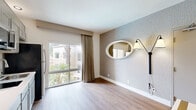 Located at 11260 Point East Dr in Rancho Cordova, California, this single-floor unit offers a thoughtfully arranged layout comprising one bedroom, two bathrooms, a kitchen, and a living room. The living area features light wood-look flooring, textured accent walls, and a large sliding glass door dressed with neutral drape curtains that invite natural light and offer views of surrounding palm trees and the neighborhood beyond. A decorative oval mirror and wall-mounted lighting add character to the space. The kitchen is efficiently designed with gray shaker-style cabinetry extending to the ceiling for ample storage, white countertops, a smooth cooktop, stainless steel appliances including a microwave and refrigerator, and a single-basin sink. An arched passageway provides an architectural detail that connects spaces with a classic touch. The spacious primary bedroom is finished with patterned carpet flooring, a wall-mounted flat-screen TV, and a large window with drape curtains that bring in soft natural light. Two bathrooms serve the unit, one featuring white tile flooring, a tub-shower combination with wood-toned tile surround, and clean white walls. The overall aesthetic is clean, neutral, and move-in ready.