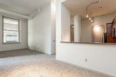 This single-floor loft-style unit combines industrial character with residential comfort throughout its thoughtfully designed layout. The open living area features impressively high ceilings with exposed structural beams and overhead ductwork, complemented by track lighting that adds a warm, focused ambiance. A half-wall with a dark granite ledge separates the living space from the kitchen, creating a natural flow between the two areas. The kitchen is well-appointed with warm wood cabinetry, granite-style countertops, hardwood flooring, and stainless steel appliances including a refrigerator with ice maker, an electric range, and an over-the-range microwave. The bedrooms are carpeted for comfort and are flooded with natural light through large windows that frame views of surrounding urban architecture. One bedroom features expansive floor-to-ceiling industrial-style windows overlooking an interior atrium, while additional bedrooms offer street-facing views. A walk-in closet with built-in hanging rods provides ample storage. The bathroom includes a dark wood vanity, a large mirror, and hardwood-style flooring. Exposed pipes and high transoms throughout lend the space a distinctive urban loft aesthetic.