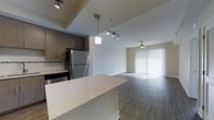 This single-floor space showcases a thoughtfully designed open-concept layout that seamlessly connects the kitchen and living area. The kitchen is equipped with stainless steel appliances, including a full-size refrigerator and dishwasher, complemented by dark wood-toned cabinetry that provides ample storage. A white quartz-style kitchen island serves as a functional centerpiece, ideal for meal preparation or casual dining. Pendant lighting above the island adds a modern touch, while a track lighting system illuminates the kitchen workspace effectively. The living area extends naturally from the kitchen, featuring durable wood-look flooring that runs throughout the space, contributing to a cohesive aesthetic. A ceiling fan in the living area enhances comfort and airflow. Vertical blinds covering a large sliding glass door or window allow for adjustable natural light, brightening the entire open floor plan. The neutral white walls and clean lines throughout give the space a contemporary and versatile ambiance, well-suited for a variety of interior styles. The overall layout prioritizes flow and functionality.