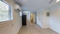 This single-floor studio is designed to maximize functionality within a well-proportioned open layout. The main living area features light-toned hardwood-style flooring that runs throughout, lending a clean, cohesive look to the space. Crisp white walls and a smooth white ceiling create a bright, airy atmosphere that enhances the sense of openness.

A wall-mounted ductless mini-split unit provides efficient heating and cooling, positioned near a window that allows natural light to filter into the space. The kitchen area is integrated into the main room, equipped with a full-size refrigerator and additional appliances visible along the wall, offering practical everyday utility without sacrificing floor space.

Toward the rear of the studio, a dedicated bathroom is accessible via a sliding barn-style door, with warm interior lighting visible from the main area. Adjacent to the bathroom entry is a full closet with bi-fold doors, providing ample storage. The open-concept design connects all functional zones seamlessly, making the studio feel organized and livable. Overall, the space presents a modern, neutral aesthetic suited to comfortable, streamlined living.