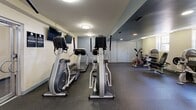 Located at 1445 Ogden St NW in Washington, DC, this fitness center offers a well-equipped, single-floor exercise space designed to support a comprehensive workout routine. The room is finished with durable rubber flooring throughout, providing a safe and functional surface for all types of exercise.

The cardio section features multiple elliptical trainers lined along one wall, each equipped with individual display consoles. Mounted flat-screen televisions are positioned for easy viewing during workouts. The strength training area is equally well-appointed, offering a selection of cable and resistance machines, adjustable benches, and a full rack of dumbbells ranging across multiple weight increments. Additional functional fitness equipment, including medicine balls and resistance bands in various sizes, rounds out the available options.

Full-length mirrors span an entire wall, visually expanding the space and allowing users to monitor their form during exercise. Natural light filters in through windows on one side of the room, complemented by flush-mounted ceiling lights that ensure bright, even illumination throughout. The layout is organized to allow clear separation between cardio and strength zones, promoting efficient use of the space. This fitness center provides residents a convenient, fully equipped venue for maintaining an active lifestyle.