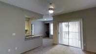 This single-floor space showcases an open and functional layout designed for comfortable everyday living. The living area is finished with soft carpeting and features a ceiling fan with integrated lighting, creating a welcoming and well-lit atmosphere. A sliding glass door with vertical blinds fills the space with natural light and provides direct access to an outdoor area, enhancing the sense of openness.

The kitchen is connected to the living area via a pass-through opening, allowing for easy interaction between the two spaces. A granite-topped breakfast bar serves as both a practical workspace and a casual dining surface. The kitchen is equipped with white cabinetry, a tile backsplash, and a stainless steel sink, offering a clean and classic aesthetic.

Beyond the living area, a secondary room is visible, featuring built-in shelving and a window that brings in additional natural light. Crown molding details along the ceiling add a refined architectural touch throughout the space.

The overall ambiance is neutral and versatile, with gray-toned walls and light carpeting providing a clean canvas. The thoughtful layout, natural light, and functional design elements make this a practical and inviting space suited for a variety of lifestyles.