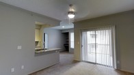 This single-floor space showcases an open and functional layout designed for comfortable everyday living. The living area is finished with soft carpeting and features a ceiling fan with integrated lighting, creating a welcoming and well-lit atmosphere. A sliding glass door with vertical blinds fills the space with natural light and provides direct access to an outdoor area, enhancing the sense of openness.

The kitchen is connected to the living area via a pass-through opening, allowing for easy interaction between the two spaces. A granite-topped breakfast bar serves as both a practical workspace and a casual dining surface. The kitchen is equipped with white cabinetry, a tile backsplash, and a stainless steel sink, offering a clean and classic aesthetic.

Beyond the living area, a secondary room is visible, featuring built-in shelving and a window that brings in additional natural light. Crown molding details along the ceiling add a refined architectural touch throughout the space.

The overall ambiance is neutral and versatile, with gray-toned walls and light carpeting providing a clean canvas. The thoughtful layout, natural light, and functional design elements make this a practical and inviting space suited for a variety of lifestyles.