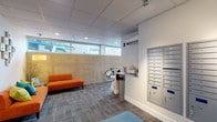 This contemporary residential lobby offers a thoughtfully designed entry experience that balances functionality with modern style. The space is laid out across a single floor and is organized into distinct zones that guide residents and guests seamlessly from entry to elevator.

The main entry corridor is bright and airy, lined with large horizontal windows that allow natural light to pour in along the length of the hallway. A textured concrete accent wall adds an industrial character, while patterned carpet tile flooring runs throughout, lending warmth and visual interest underfoot. A bicycle storage area and a convenient amenity station with umbrella storage and informational brochures are tucked neatly along the corridor.

The lounge area is a vibrant focal point, furnished with bold orange sofas accented by colorful throw pillows in teal and green. A warm floor lamp, decorative wall art, and a side table complete the inviting seating nook, which sits adjacent to the elevator bank framed by a distinctive wood mosaic tile accent wall.

The mailroom area is both practical and well-appointed, featuring an extensive array of individual metal mailboxes covering a full wall, served by track lighting overhead. A teal upholstered ottoman provides a convenient perch. The drop ceiling with recessed and track lighting maintains a clean, polished aesthetic throughout the entire lobby.