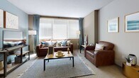 This single-floor unit is thoughtfully laid out to maximize shared and private living spaces. The open-concept living and dining area forms the heart of the unit, where a generously sized window dressed with blue curtains and horizontal blinds fills the space with natural light. Brown leather seating, a central coffee table, and a patterned area rug create a comfortable, casual atmosphere, while a wall-mounted television completes the lounge setup.

The kitchen is well-equipped with dark espresso cabinetry, granite-style countertops, and stainless steel appliances including a refrigerator, electric range, microwave, and dishwasher. Wood-look flooring and a round dining table with seating provide a practical everyday dining area directly adjacent to the kitchen.

The bedrooms feature carpeted floors, natural light from windows with blinds and drape panels, and dedicated study areas with shelving and desk setups — well-suited for student living. The unit includes two bathrooms: one with a dark vanity cabinet, large mirror, and wood-look flooring, and another featuring a full bathtub with a decorative shower curtain and accent blue walls. Overall, the unit offers a functional, organized environment suited for shared occupancy.