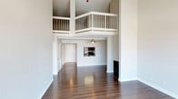 This two-story space is defined by its dramatic vertical scale, with soaring ceilings that draw the eye upward to a loft level overlooking the main living area below. A white-railed balcony runs along the upper floor, creating an open, airy connection between the two levels and adding architectural interest to the space.

The main living area features rich, dark hardwood flooring that extends throughout, providing a warm contrast to the crisp white walls and trim. A fireplace anchors one side of the room, adding a functional and aesthetic focal point. A ceiling fan is installed in an adjoining area, indicating a secondary zone that flows naturally from the main living space.

The kitchen is visible from the living area and includes cabinetry and black appliances, maintaining a clean, functional aesthetic consistent with the rest of the space. Additional doorways lead to further rooms, suggesting a well-connected floor plan.

The overall ambiance is clean, open, and modern, with the interplay of high ceilings, reflective flooring, and the loft overlook giving the space a notably expansive and architecturally distinctive character.