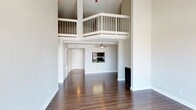 This two-story space is defined by its dramatic vertical scale, with soaring ceilings that draw the eye upward to a loft level overlooking the main living area below. A white-railed balcony runs along the upper floor, creating an open, airy connection between the two levels and adding architectural interest to the space.

The main living area features rich, dark hardwood flooring that extends throughout, providing a warm contrast to the crisp white walls and trim. A fireplace anchors one side of the room, adding a functional and aesthetic focal point. A ceiling fan is installed in an adjoining area, indicating a secondary zone that flows naturally from the main living space.

The kitchen is visible from the living area and includes cabinetry and black appliances, maintaining a clean, functional aesthetic consistent with the rest of the space. Additional doorways lead to further rooms, suggesting a well-connected floor plan.

The overall ambiance is clean, open, and modern, with the interplay of high ceilings, reflective flooring, and the loft overlook giving the space a notably expansive and architecturally distinctive character.