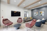 This thoughtfully designed amenity space spans a generous single floor, offering a seamless blend of social, recreational, and functional zones. The lounge area anchors one end of the space, showcasing exposed natural wood ceiling beams contrasted against crisp white painted brick walls. Contemporary furnishings — including sculptural burgundy leather chairs, a neutral sectional sofa with yellow accent pillows, and a sleek wall-mounted flat-screen TV — create an inviting, curated atmosphere. Bold abstract artwork adorns a deep charcoal feature wall, adding a gallery-like quality to the seating nook.

A striking central wet bar serves as the architectural centerpiece, featuring dark cabinetry with brass hardware, a stone countertop, a built-in wine refrigerator, and a bold geometric black-and-white tile floor that commands attention. Flanking perimeter counters with matching cabinetry and large industrial-style windows allow natural light to pour in.

The game and dining room offers wall-mounted television display, a foosball table, and casual dining tables with warm tan leather chairs set against charcoal-painted walls. A fully equipped catering kitchen — outfitted with wood-grain cabinetry, gold hardware, white quartz countertops, checkered tile flooring, and stainless steel appliances — rounds out the space with practical elegance throughout.