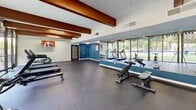 Located at 228 Morrow Rd in Forest Park, Georgia, this fitness center offers a thoughtfully designed exercise space spread across a single open floor. The room is distinguished by bold, warm-toned exposed structural beams that contrast beautifully against the white ceiling, lending an industrial yet inviting character to the space.

Abundant natural light pours in through expansive floor-to-ceiling windows that line multiple walls, providing sweeping views of lush greenery, manicured lawns, and a nearby outdoor pool area. The large mirror panels along one wall further amplify the sense of openness and allow users to monitor their form during workouts.

The fitness center is comprehensively equipped to support a variety of training styles. Cardio options include treadmills and an elliptical trainer, while a spin bike and rowing machine cater to low-impact, high-intensity workouts. Strength training is well-represented with adjustable benches, a punching bag, medicine balls, kettlebells, and additional free weights arranged on a dedicated rack. A wall-mounted television adds entertainment for those on cardio machines.

The flooring throughout is a durable dark rubber material, practical for both heavy equipment and dynamic movement. The teal accent walls add a vibrant pop of color, complementing the overall modern aesthetic. This fitness center delivers a complete and energizing environment for a wide range of workout routines.
