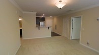 This single-floor residence offers a thoughtfully designed open-concept layout that seamlessly connects the living area to the kitchen. The living space is finished with plush neutral-toned carpeting, crown molding, and a classic pendant light fixture that anchors the room with warm, ambient illumination. Recessed lighting further brightens the interior, creating a well-lit environment throughout.

The kitchen is defined by a raised breakfast bar with seating, providing a natural divide between the cooking and living areas. Dark espresso-toned cabinetry provides a rich contrast against the light countertops, while stainless steel appliances add a contemporary touch. The kitchen floor transitions to a darker hard surface, distinguishing it from the carpeted living space.

Crown molding runs consistently along the ceiling, lending the space an added sense of polish and architectural detail. A utility or electrical panel is visibly integrated into the wall near the entryway, and a set of paneled doors leads to additional rooms or storage areas. The neutral wall tones throughout contribute to a clean, versatile aesthetic that suits a range of furnishing styles. Overall, the space presents a functional and well-appointed single-level layout with practical finishes and classic design elements.