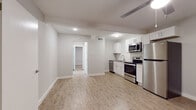 This single-floor unit showcases a thoughtfully updated interior with a cohesive, modern design throughout. Light wood-style luxury vinyl plank flooring runs continuously across all rooms, creating a warm and unified feel. The open kitchen is well-appointed with white shaker-style cabinetry, grey countertops, and a full suite of stainless steel appliances including a range, over-the-range microwave, dishwasher, and refrigerator. A white subway tile backsplash adds a clean, classic detail to the cooking area, which flows directly into an open living space with a ceiling fan overhead.

A hallway leads to the additional rooms, each finished with the same light flooring and neutral grey walls. One bedroom features a ceiling fan with a light fixture and a window that brings in natural light. A second room offers a versatile, open layout suitable for various uses. The bathroom is finished with large-format marble-style tile lining the tub surround, complemented by wood-look flooring, a white vanity, and brushed metal hardware. Throughout the unit, recessed lighting and clean white trim contribute to a bright, well-maintained atmosphere.